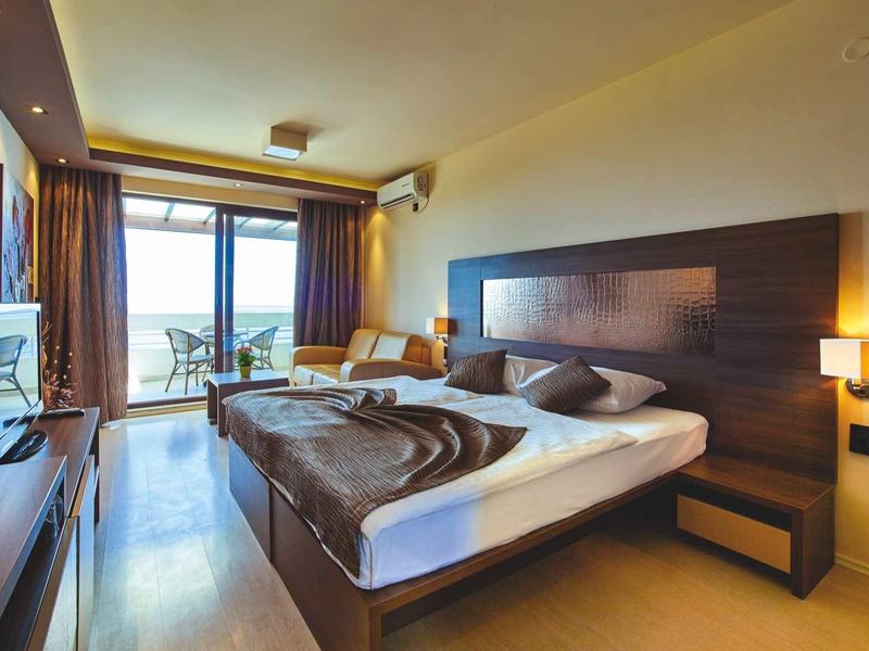 Modern hotel room with double bed, balcony, and sea view in daylight.