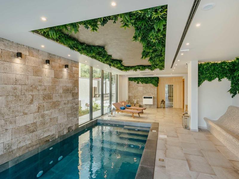 Piscine intérieure moderne avec carrelage en mosaïque, plantes vertes au plafond et murs en pierre naturelle.