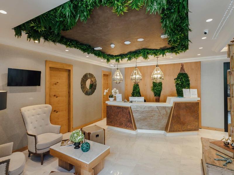 Hall d'hôtel moderne avec réception, fauteuils confortables, plantes vertes et plafond en bois décoratif.