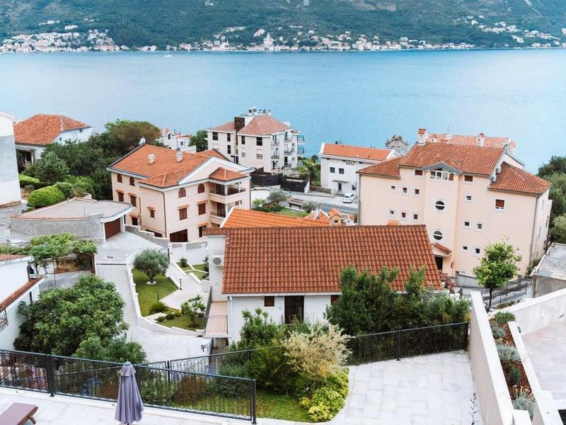 Vue d'un village côtier avec des toits rouges et une piscine devant les montagnes et la mer.