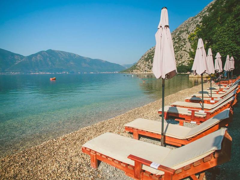 Sillas de playa con sombrillas alineadas en una costa rocosa con montañas al fondo.