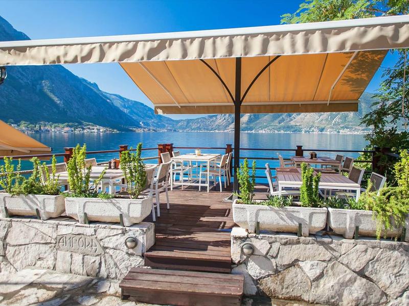 Terraza con sillas y mesas frente a un lago con montañas al fondo bajo cielo azul.
