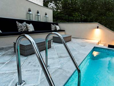 Moderner Poolbereich mit Mosaikfliesen, Edelstahlleiter, Sitzbank mit Kissen und stimmungsvollem Licht.