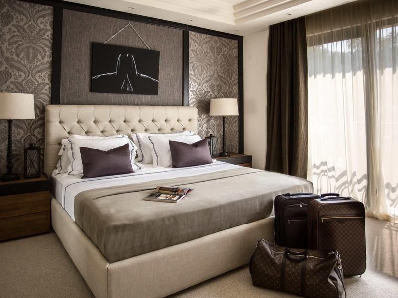 Modernes Schlafzimmer mit großem gepolstertem Bett, zwei Nachttischen, Lampen und großen Fenstern.