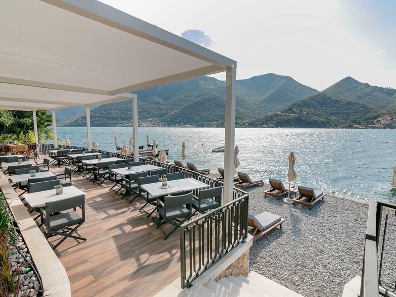 Terrassenrestaurant mit weißen Tischen und Stühlen neben ruhig blauem Meer und Bergen im Hintergrund.