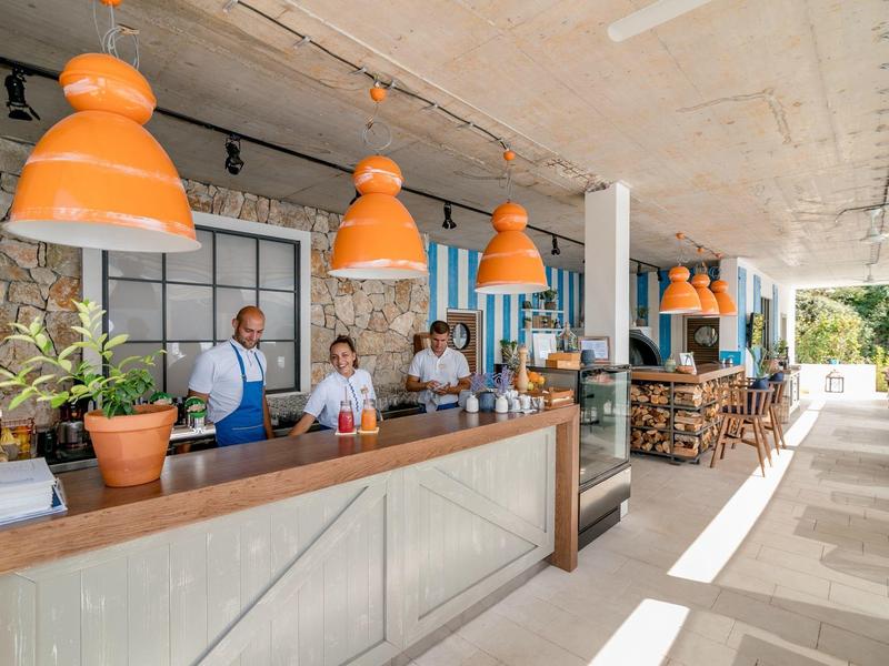 Moderner Außenbereich einer Bar mit orangenen Lampen, Holzmöbeln und grünen Pflanzen am Tresen.