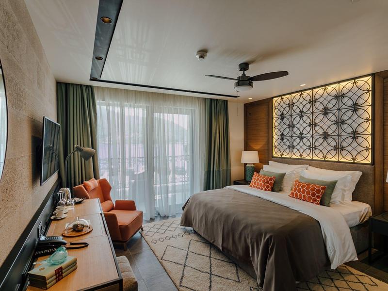 Modernes Hotelzimmer mit großem Bett, gemütlichem Sessel und hellem Tageslicht durch Fenster.