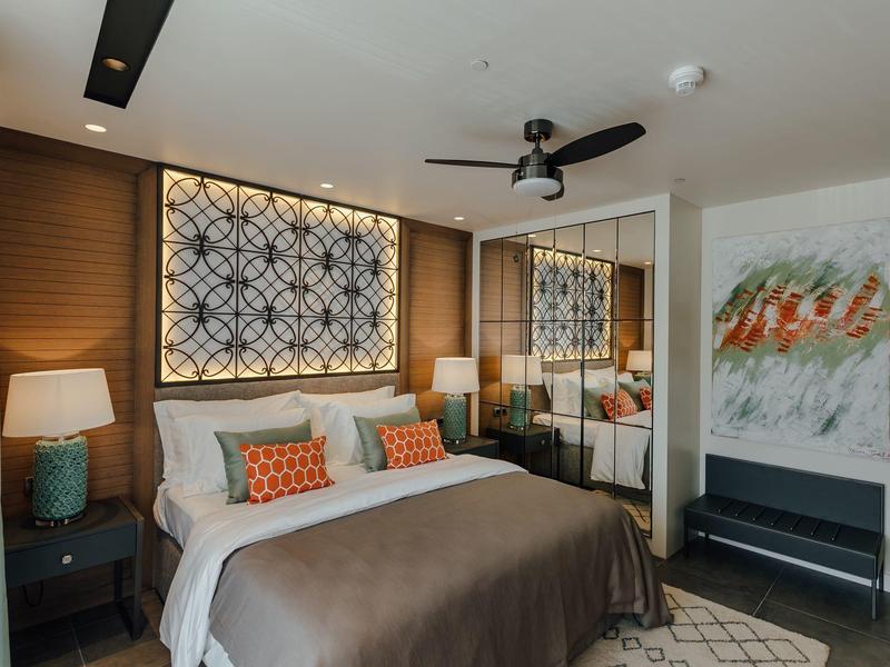 Modernes Schlafzimmer mit großem Bett, bunten Kissen, Lampen, Deckenventilator und Spiegelwand.