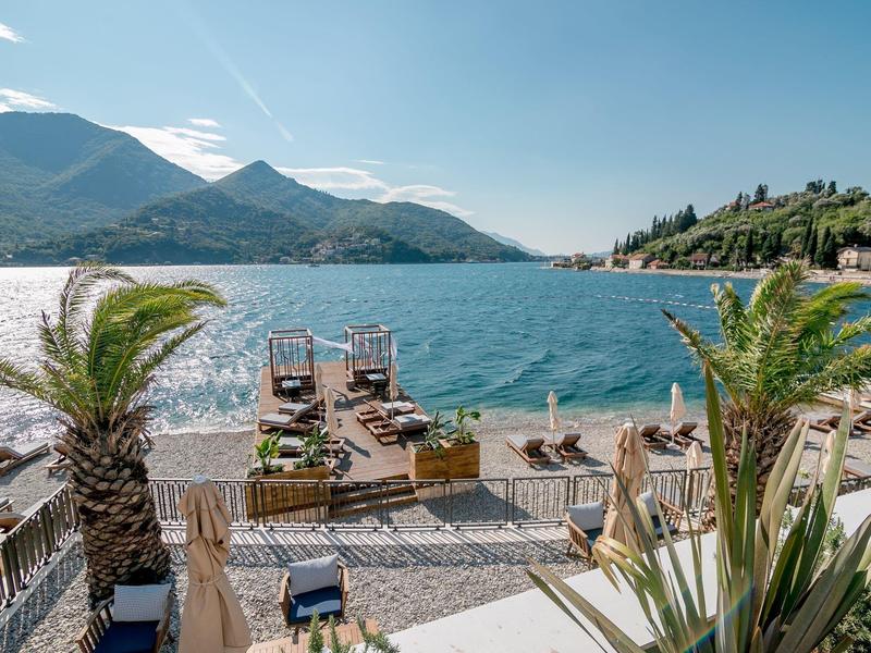 See mit Steg und Liegestühlen, umgeben von Bergen und Palmen unter blauem Himmel.