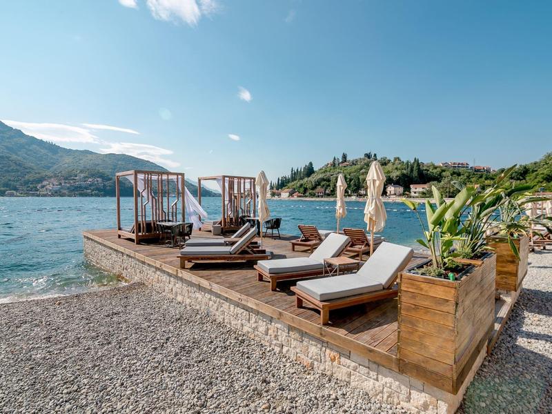 Strandbereich mit Holzterrasse, Liegen, Sonnenschirmen und Blick auf blaues Wasser und grüne Hügel.