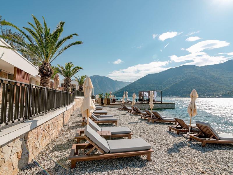 Sonnenliegen am Kiesstrand neben Palmen, Blick auf Berge und Sonne über dem Wasser.