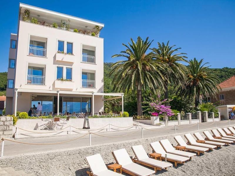 Modernes weißes Gebäude mit Balkonen, davor Palmen und Liegestühle auf kieseligen Strand.