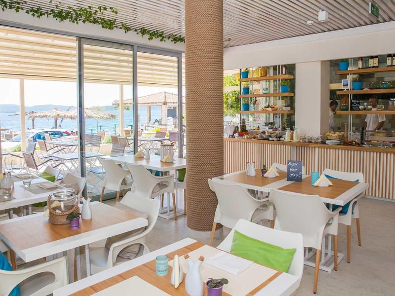 Modernes Café mit weißen Tischen, bunten Kissen und großer Glasfront mit Meeresblick.