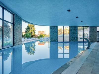 Innenraum mit großem, blau beleuchtetem Schwimmbad und bodentiefen Fenstern zum Außenbereich.