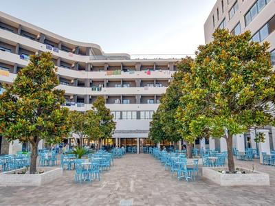 Freier Platz mit blauen Stühlen, großen Bäumen und modernen Gebäuden im Hintergrund.