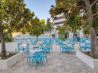 Leerer Außenbereich eines Cafés mit blauen Stühlen und Tischen, umgeben von Bäumen und Steinboden.