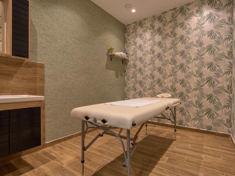 Einfach eingerichteter Behandlungsraum mit Massageliege, Holzfußboden und gemusterter Tapete.