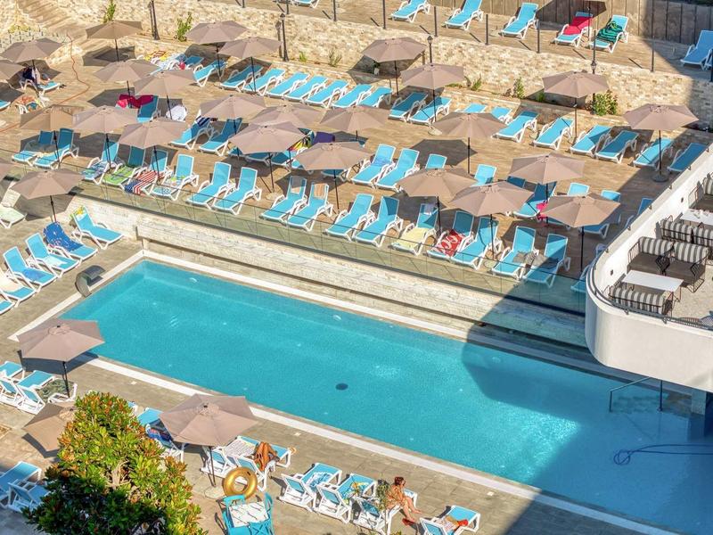 Großer Pool mit blauer Wasserfläche, umgeben von vielen blauen Liegestühlen und Schirmen.