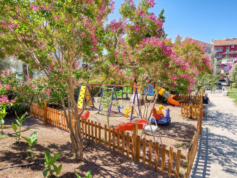 Kleiner Spielplatz mit Sand, Schaukel, Rutsche, Bänken und blühenden Bäumen umgeben von Zaun.