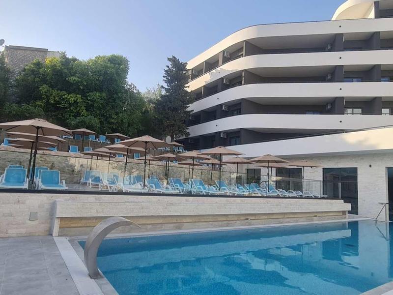 Moderner Hotelpool mit blauer Umrandung, Liegestühlen und Sonnenschirmen neben mehrstöckigem Gebäude.