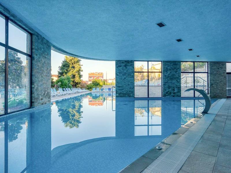 Innenraum mit großem, blau beleuchtetem Schwimmbad und bodentiefen Fenstern zum Außenbereich.