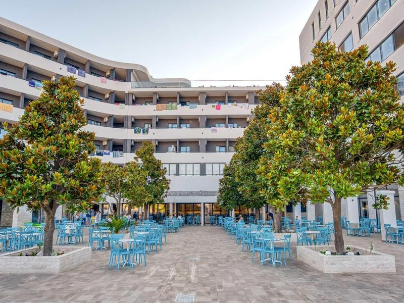 Freier Platz mit blauen Stühlen, großen Bäumen und modernen Gebäuden im Hintergrund.