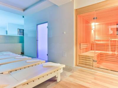 Innenraum mit Holzliegen und einer Glastür zur rot beleuchteten Sauna im Wellnessbereich.