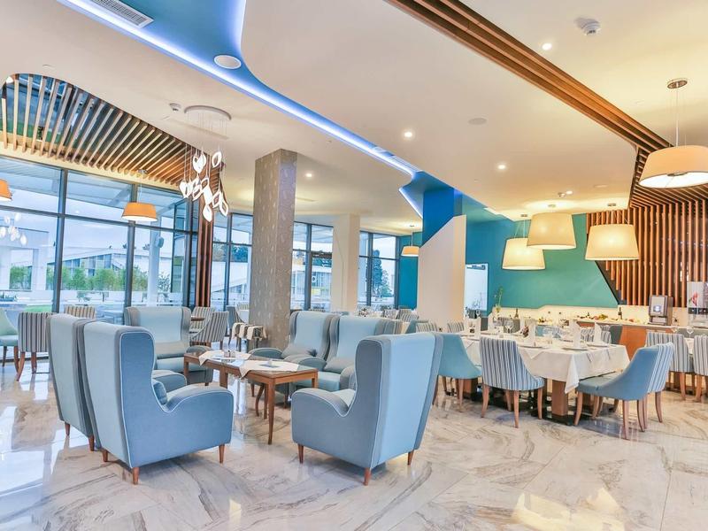 Moderne, helle Restaurantlounge mit blauen Sesseln, Holzelementen, großen Fenstern und stilvoller Beleuchtung.