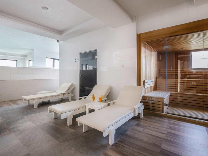 Ruheraum mit weißen Liegestühlen und angrenzender Sauna aus Holz und Glas.