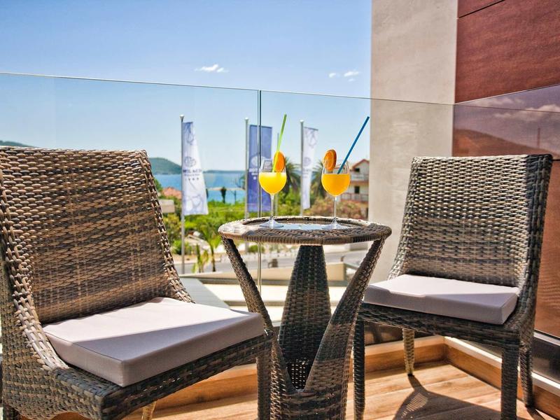 Balkon mit Tisch und zwei Korbstühlen, drei bunte Cocktails, blauer Himmel und Stadtblick.