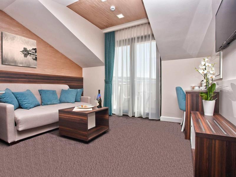 Gemütliches Zimmer mit Sofa, Kissen, Holzmöbeln, Teppichboden und großem Fenster mit Vorhängen.