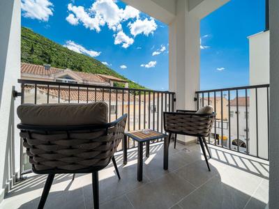 Balkon mit zwei Korbstühlen und kleinem Tisch, Blick auf Dächer und bewaldeten Hügel bei blauem Himmel.