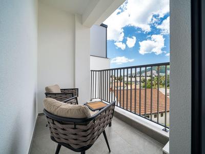 Malý balkon se dvěma polstrovanými židlemi s výhledem na střechy a modrou oblohu.