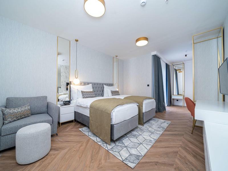 Helles, modernes Schlafzimmer mit Doppelbett, Teppich, Sofa, Fernseher und Schreibtisch in neutralen Farben.