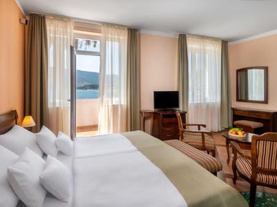 Helles Hotelzimmer mit zwei Betten, zwei Stühlen am Tisch, Fernseher und Balkon mit Ausblick.