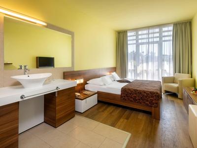 Modernes Hotelzimmer mit Doppelbett, Waschbecken, Fernseher und großen Fenstern mit Vorhängen.