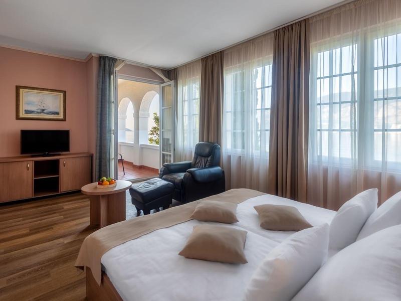 Helles Hotelzimmer mit Doppelbett, Sesseln, TV und Zugang zu Balkon mit Blick auf Meer.