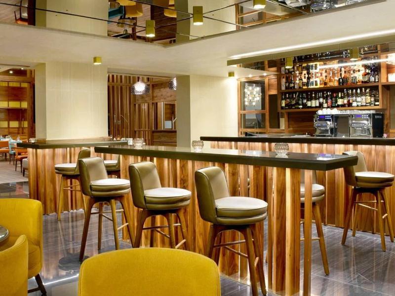 Moderne Bar mit Holztheke, gepolsterten Barhockern und gelben Sesseln, warme Beleuchtung.