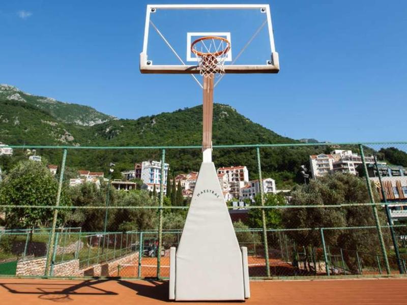 Basketballkorb auf rotem Spielfeld mit Berglandschaft und Gebäuden im Hintergrund.