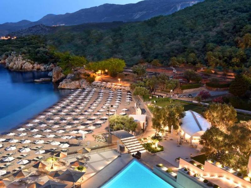 Strand mit vielen weißen Schirmen, dahinter grüne bewaldete Hügel und beleuchtete Gebäude.