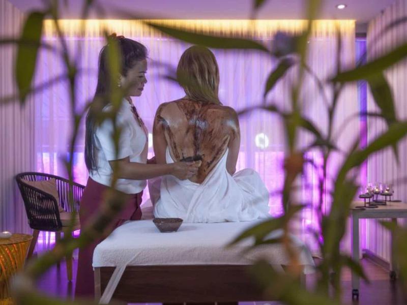 Zwei Frauen im warm beleuchteten Raum, eine sitzt auf Massagetisch, die andere berührt ihren Rücken.