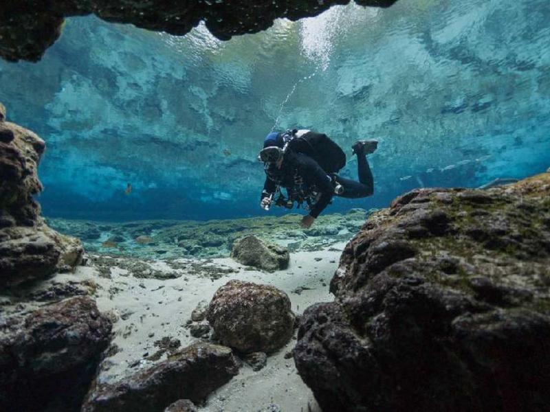 Taucher erkundet unter Wasser eine felsige und sandige Meereslandschaft, Blau- und Brauntöne dominant.