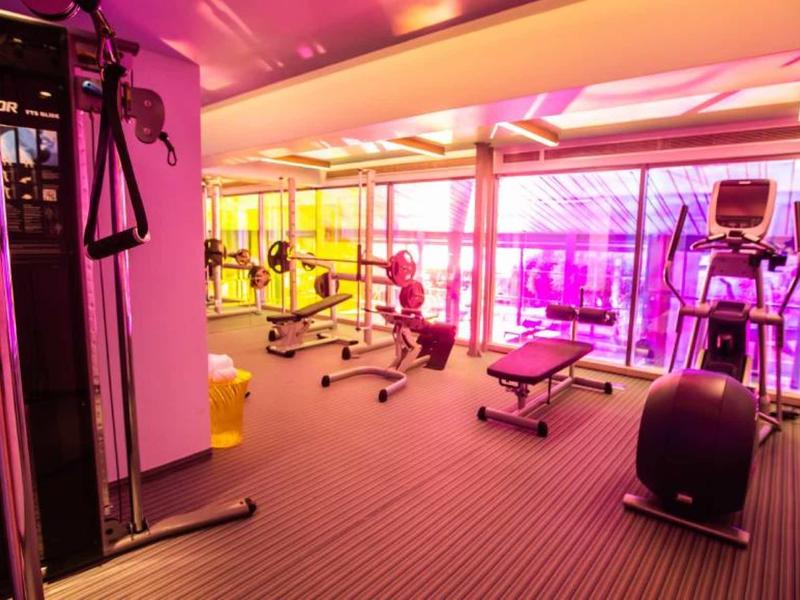 Helles Fitnessstudio mit verschiedenen Trainingsgeräten, großen Fenstern und farbigem Licht.