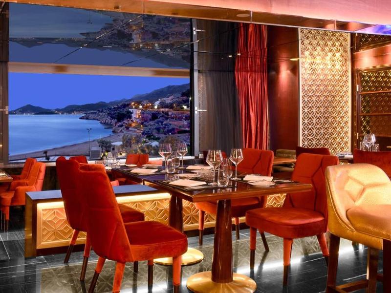 Elegantes Restaurant mit rotem Mobiliar und großem Fenster mit Blick auf Küstenlandschaft bei Nacht.