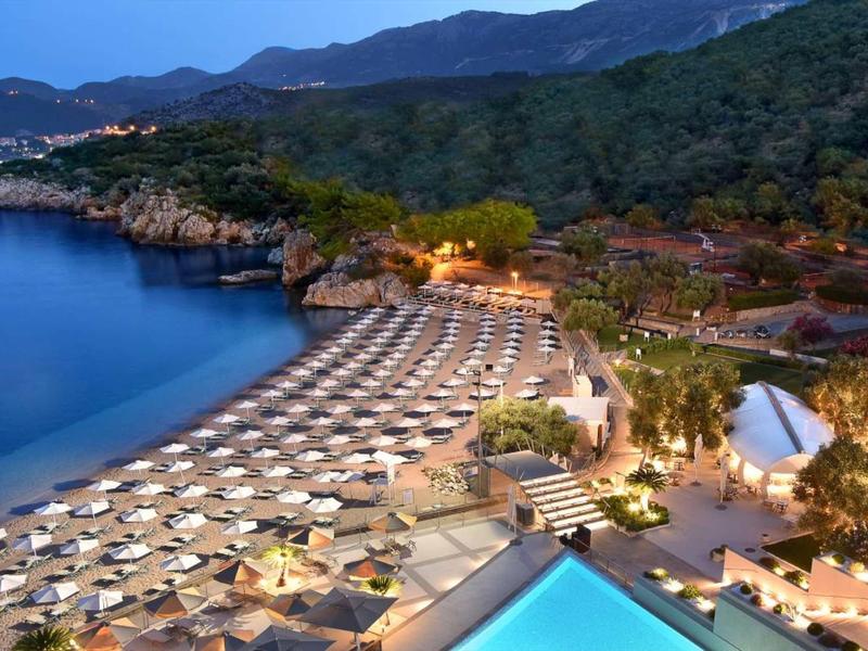 Strand mit weißen Sonnenschirmen, blauem Pool und bewaldeten Hügeln im Abendlicht.