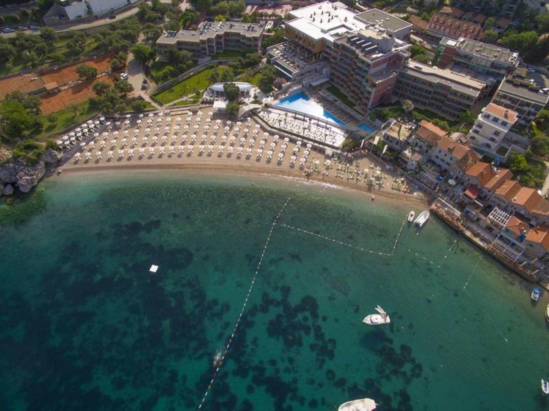 Luftaufnahme eines Strandes mit klares blaues Wasser, Liegen, Sonnenschirmen und umliegenden Gebäuden.