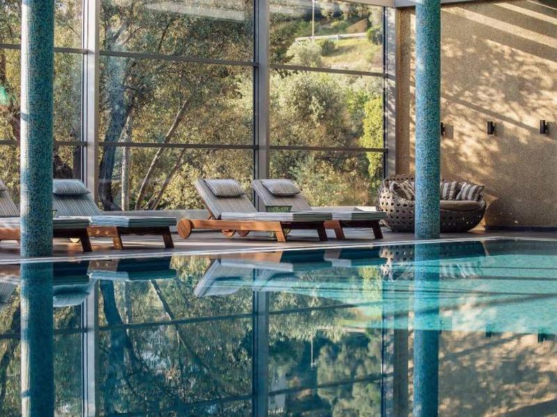 Innenraum mit klarem Schwimmbecken, Liegestühlen und großen Fenstern mit Blick auf Natur.