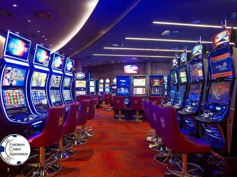 Eine lange Reihe bunter Spielautomaten mit roten Hockern in einem Casino mit rotem Teppich.