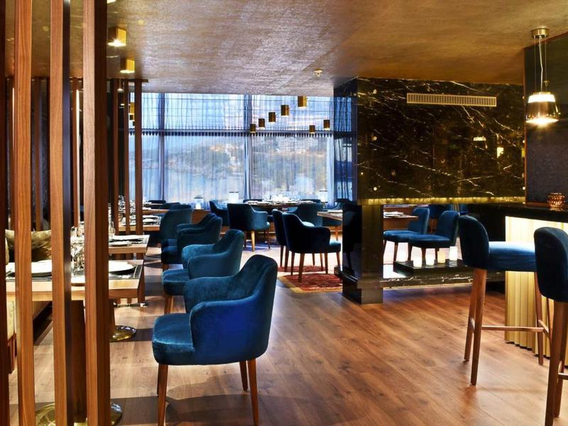 Moderner Restaurantraum mit blauen Sesseln, Holzboden und großen Fenstern für Tageslicht.