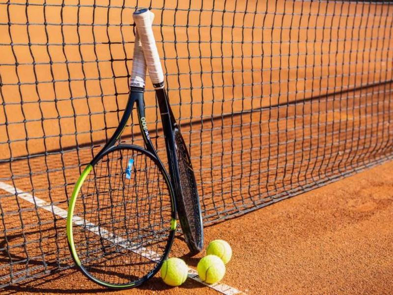 Zwei Tennisschläger lehnen am Netz, davor liegen drei gelbe Tennisbälle auf dem roten Sandplatz.